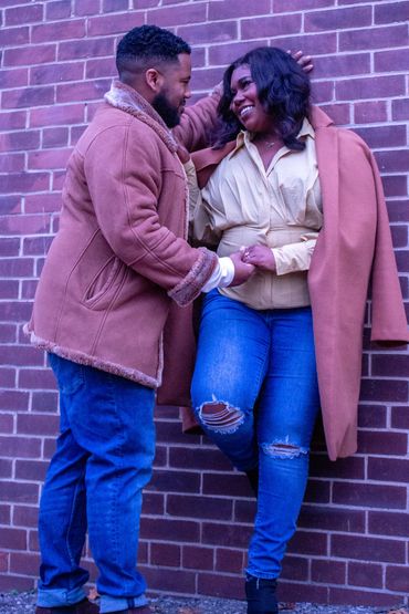 Couple smiling warmly at each other against a brick wall.