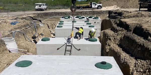 Workers install large concrete tanks in a construction trench under a clear sky.