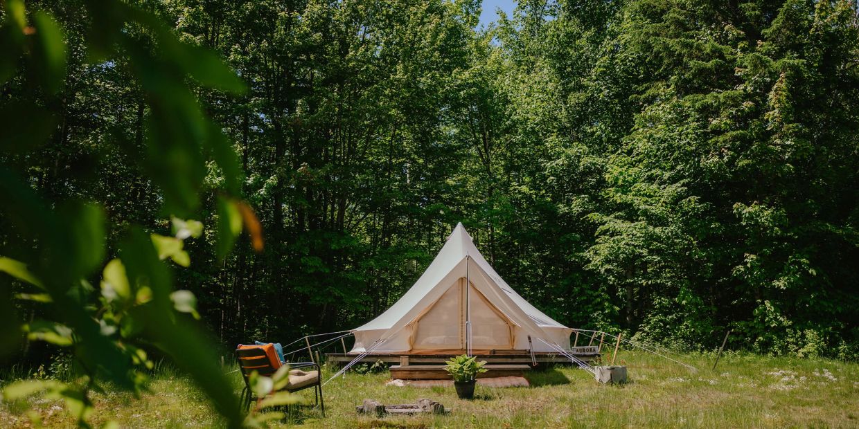 A white bell tent set up in a green forest clearing with a chair and potted plant.