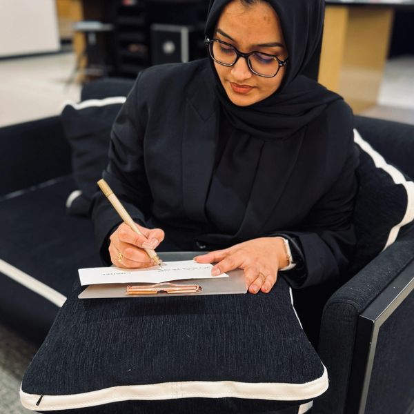 Woman in a hijab writing on paper with a pen while seated on a black couch.