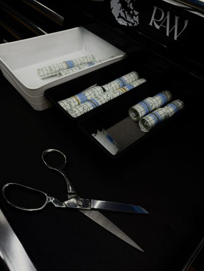 Metal scissors and rolled RAW papers in trays on a black surface.