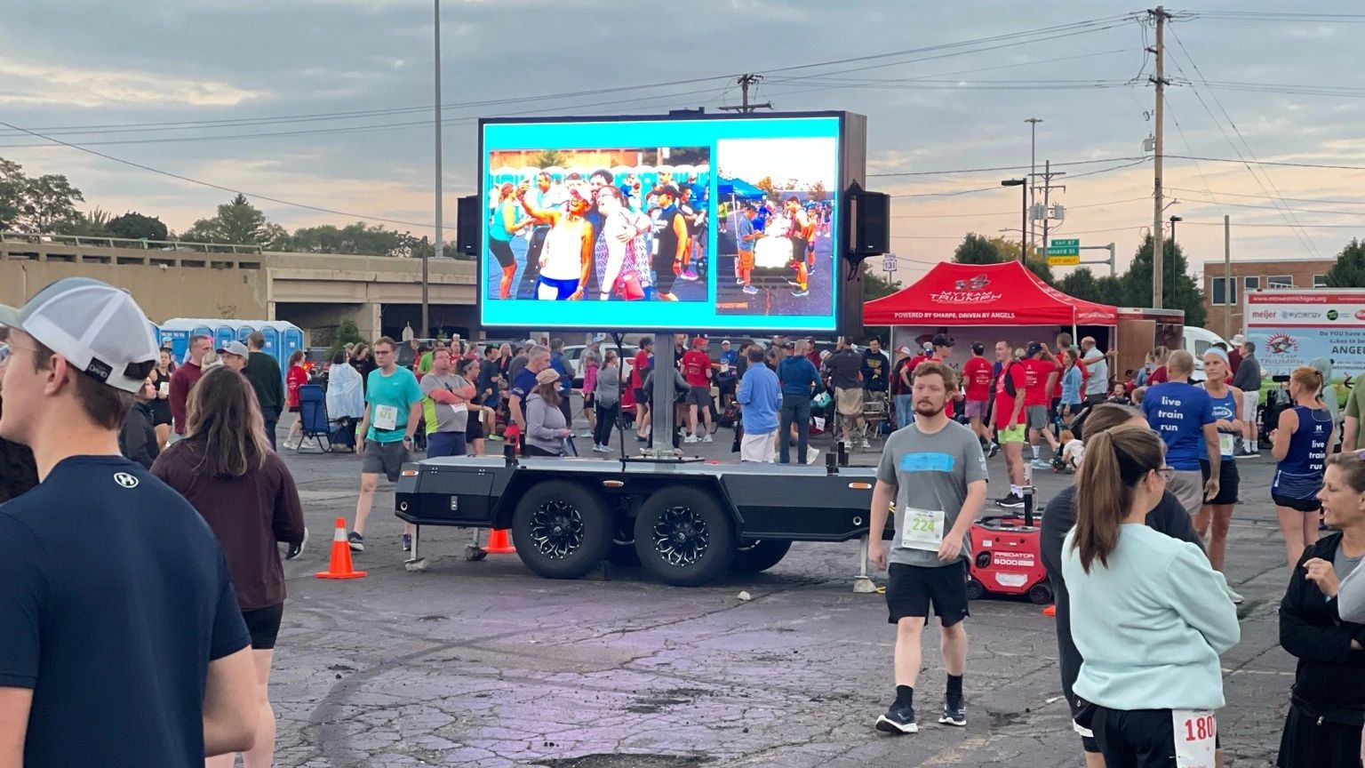 Crowd gathered at an outdoor event with a large screen and people in athletic wear.