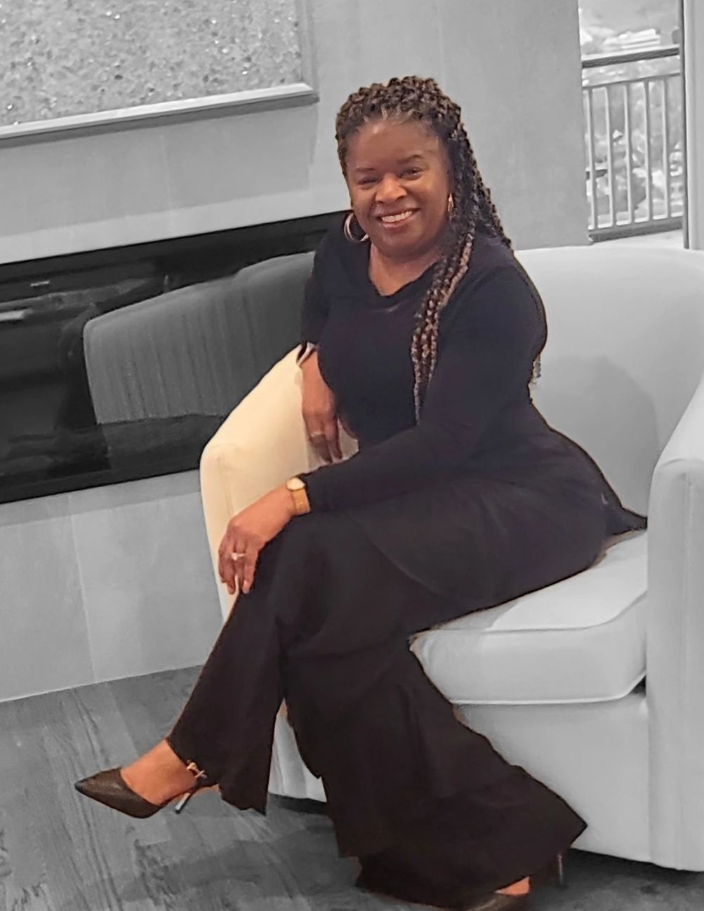 Woman in black outfit seated on a white chair, smiling warmly.