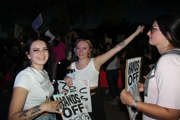Three women at a nighttime protest holding "Hands Off" signs.