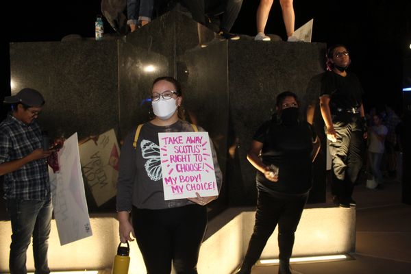 People protesting with signs at night, one with a mask holds a sign on reproductive rights.