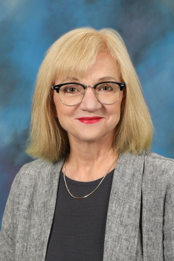Smiling woman with glasses and blonde hair wearing a grey blazer.