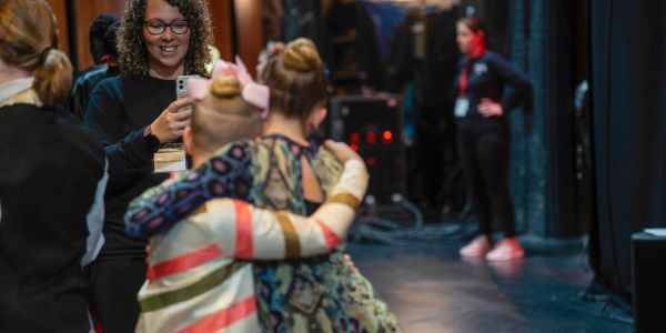 Woman taking photo of two hugging girls backstage.
