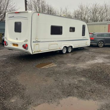 White caravan trailer parked on gravel near cars and trees.