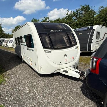 A white Swift Sprite caravan hitched to a blue vehicle under a partly cloudy sky.