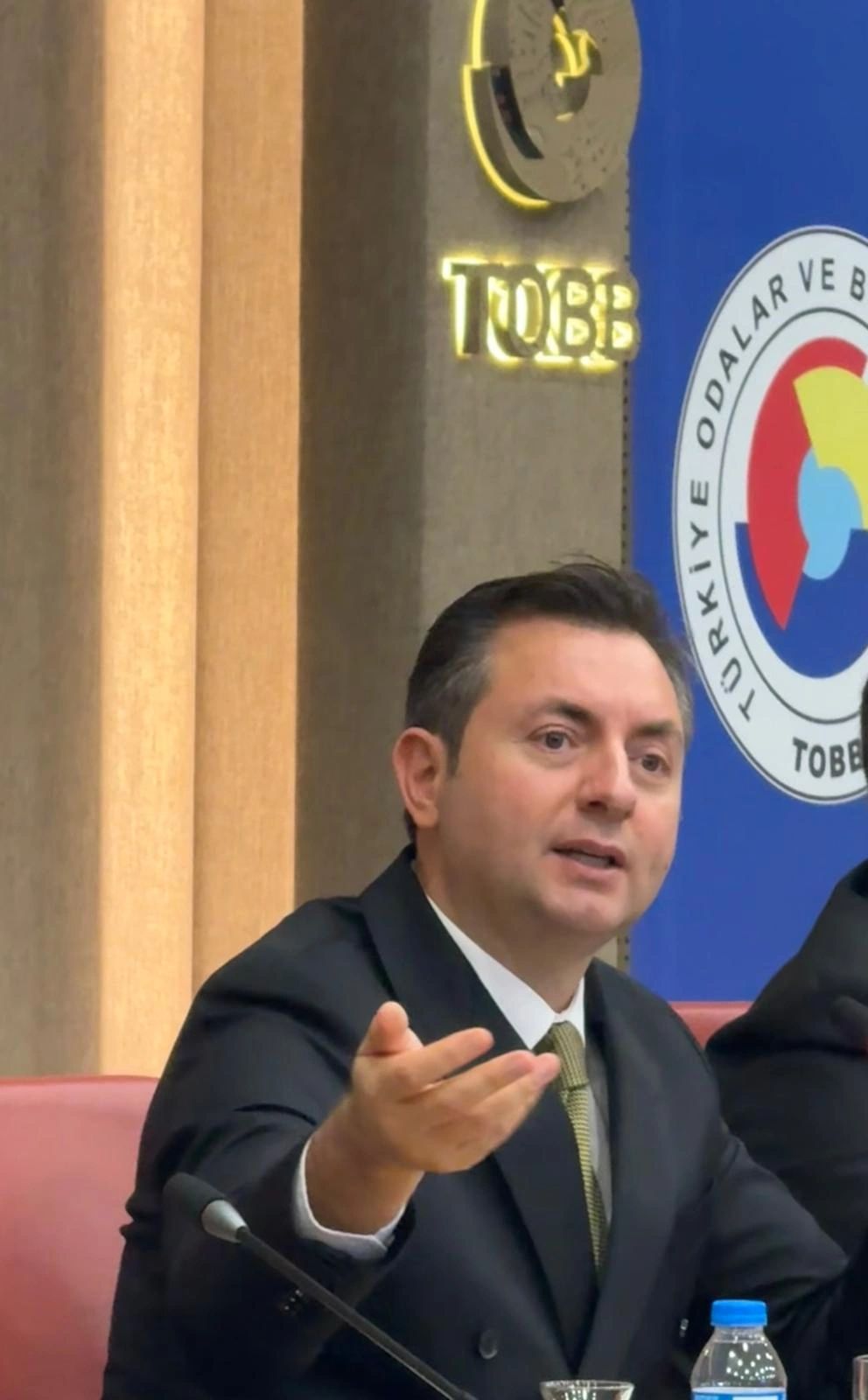 A man in a suit speaking at a conference with a microphone and water bottle.
