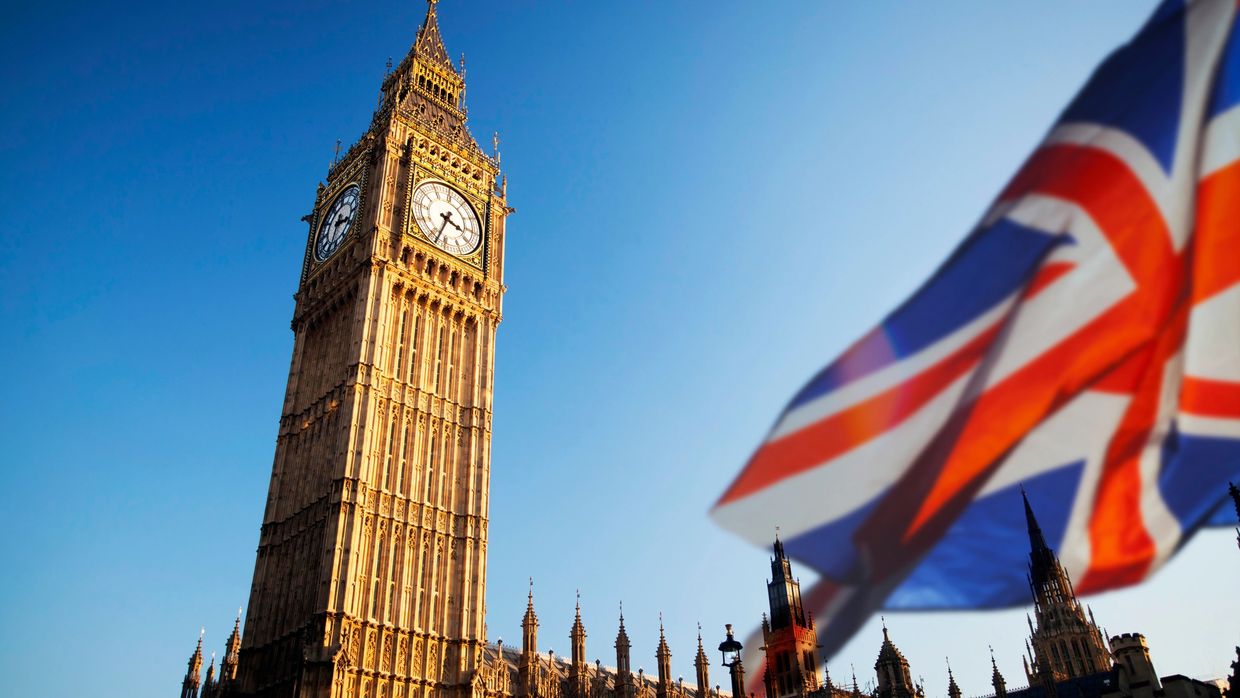 Big Ben clock tower with a waving UK flag in the foreground under a clear blue sky.