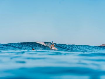 A surfer rides a wave on a clear blue ocean under a bright sky.
