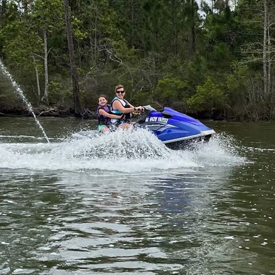 Two people riding a blue jet ski on a lake surrounded by trees.