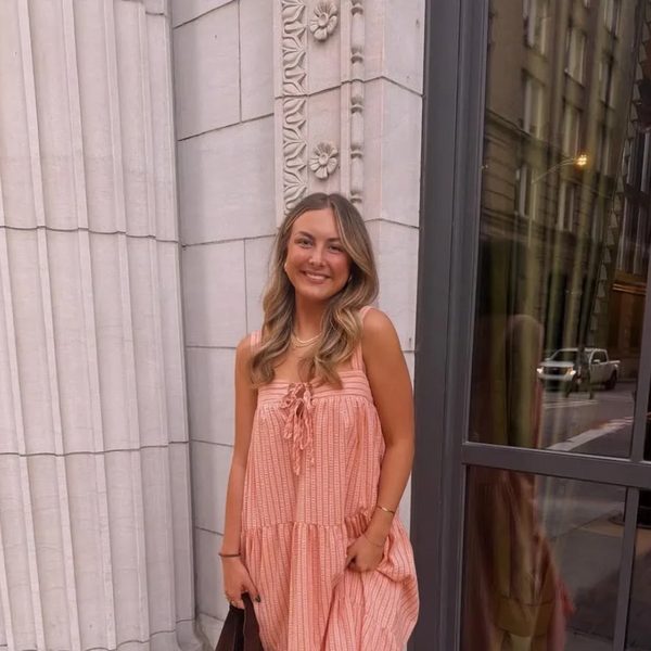 Young woman in a peach dress smiles by an ornate stone wall and glass door in the city.
