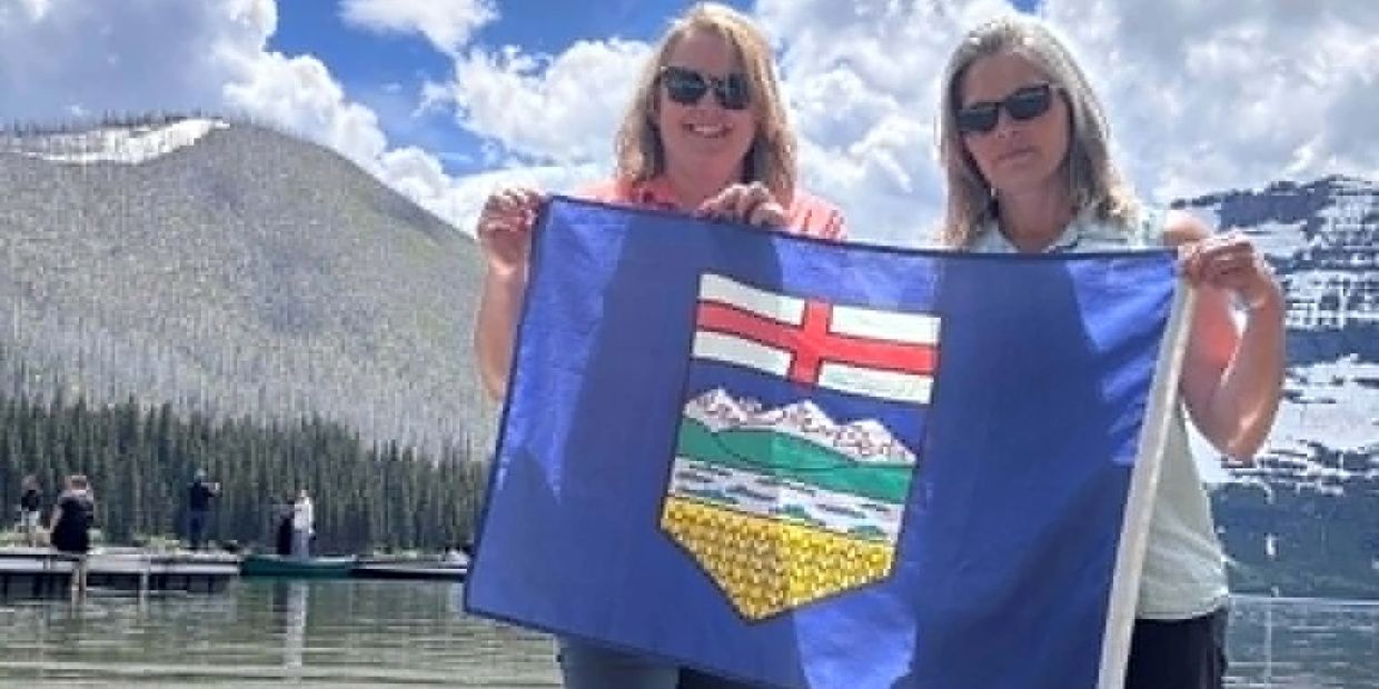 Two women holding an Alberta flag by a mountain lake.