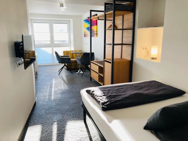 Bright, modern studio apartment with a bed, seating area, and colorful wall art.