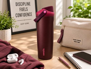 A maroon travel mug on a wooden table surrounded by accessories.
