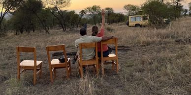 Couple sitting on wooden chairs in a savannah at sunset, with a safari vehicle nearby.