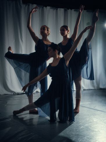 Three ballerinas in dark dresses gracefully pose in a dimly lit studio.