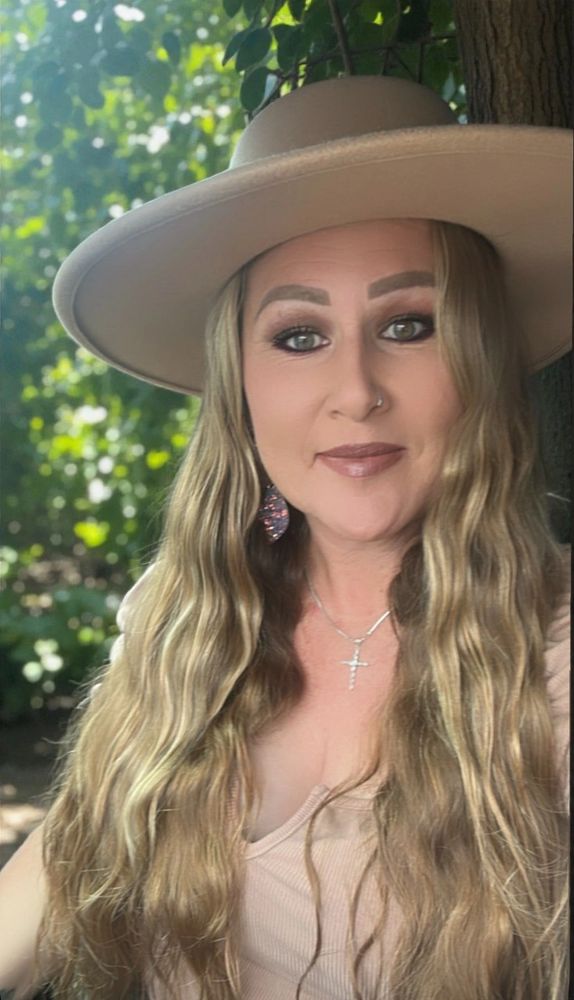 Woman with long wavy blonde hair, wearing a beige hat and necklace, smiling outdoors.