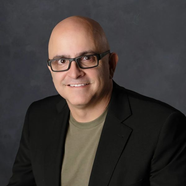 Smiling bald man wearing glasses and a black blazer against a dark background.