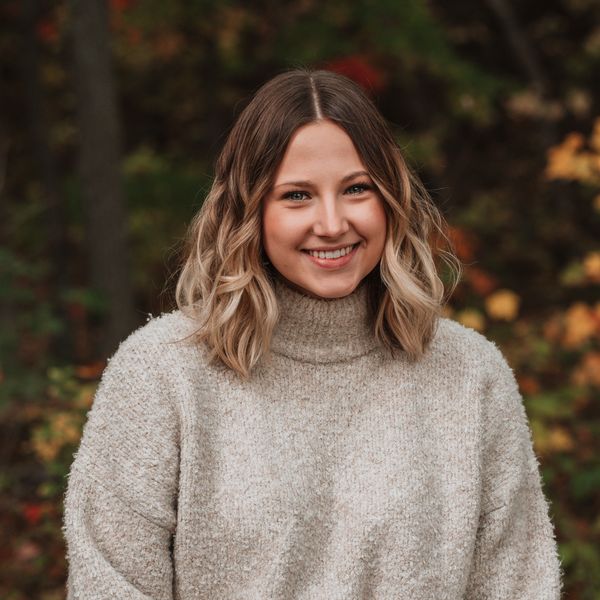 Smiling woman in cozy beige sweater outdoors with autumn foliage.