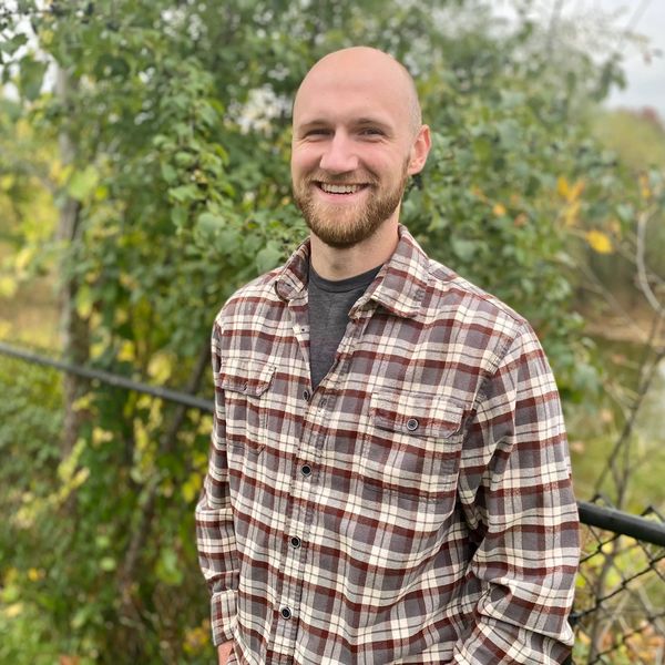 Smiling man in a plaid shirt outdoors with greenery background.