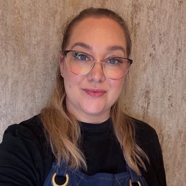A woman with glasses wearing a blue apron and black shirt smiles gently.