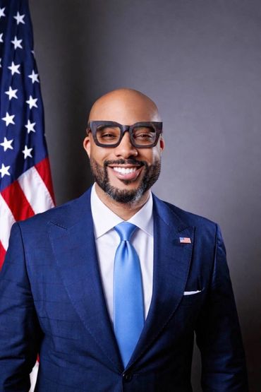 Smiling man in blue suit with American flag background.