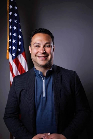 Smiling man in a blazer stands before an American flag.