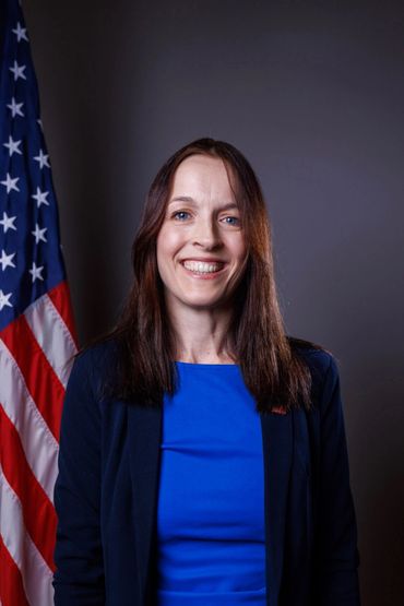 Smiling woman in blue with American flag behind her.