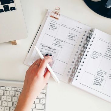 Person writing in a planner next to a keyboard and laptop.