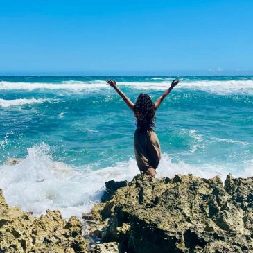 Woman standing on rocky shore with arms raised facing the ocean.