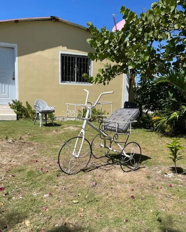 Decorative white metal bicycle planter with a cushion in a garden by a yellow house.