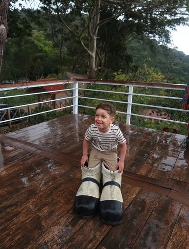 A smiling boy wearing oversized boots on a wooden deck outdoors.