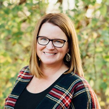 Co-founder Sarah Gleason. Woman in black shirt with plaid sweater smiling at the camera.