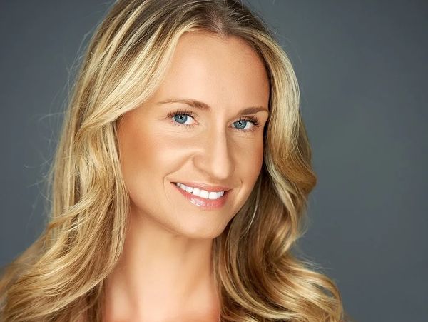 Smiling woman with long blonde hair and blue eyes against a gray background.