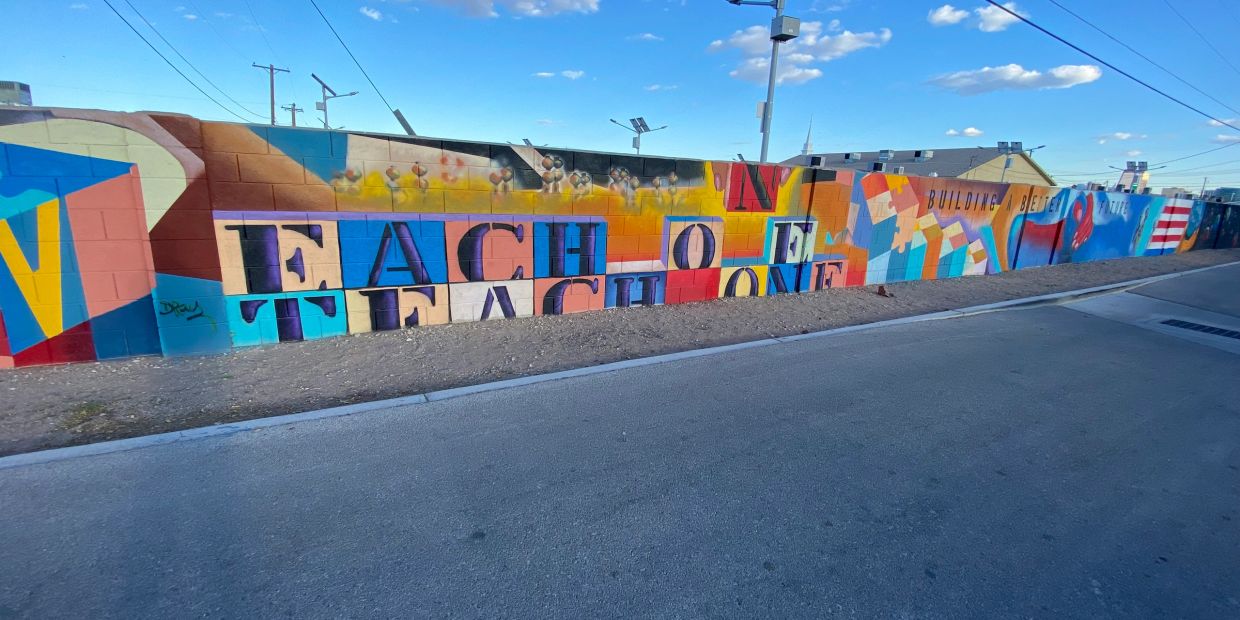 Colorful mural with the phrase "Each One Teach One" on a long wall under a clear blue sky.