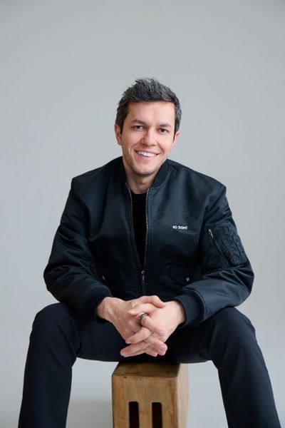 Smiling man in a black jacket sitting on a wooden stool against a plain background.
