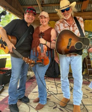 Three smiling musicians holding instruments on a wooden porch.
