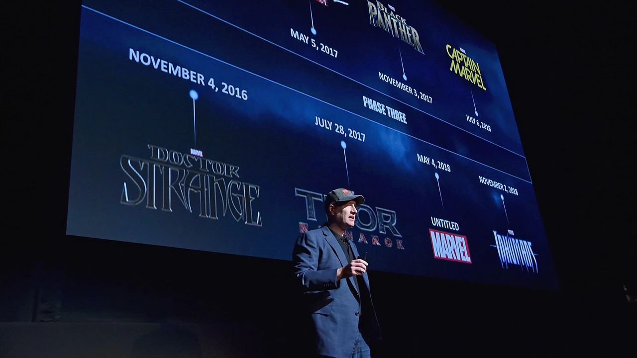 A man speaking in front of a large screen displaying a timeline of Marvel movie releases, with titles and dates for various films.