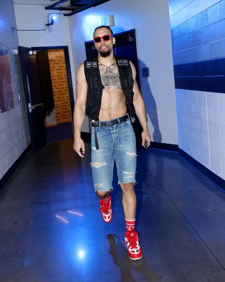 A man confidently walks through a hallway, wearing a sleeveless top, distressed denim shorts, and red sneakers. He has sunglasses on and displays several tattoos on his chest and arms.