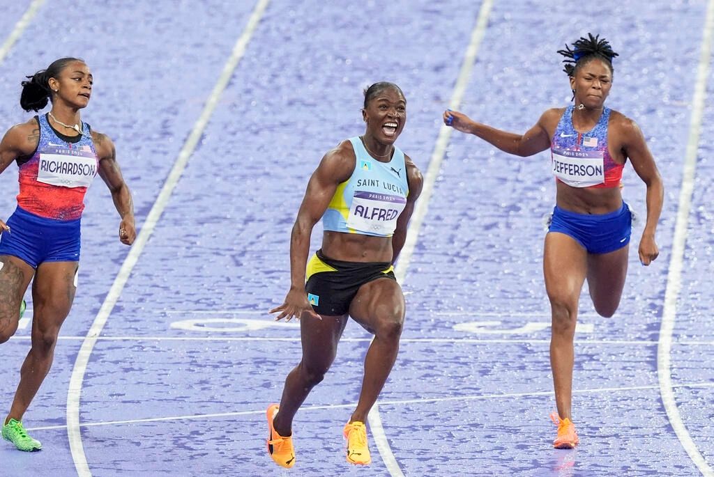 Julien Alfred celebrating her victory in the 100-meter dash, with Sha'Carri Richardson and another competitor in the background.