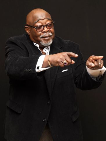 A joyful man in a black suit pointing with both hands against a black background.