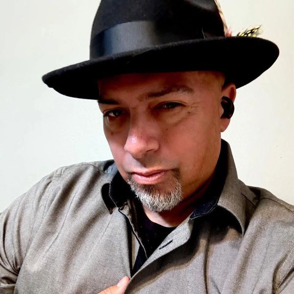 Man wearing a black fedora and gray shirt with a confident expression.