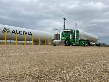 Green and white tanker truck parked near large fuel storage tanks.