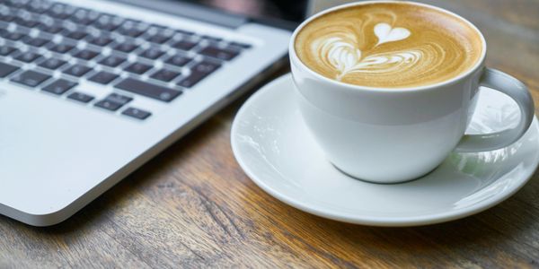 A cup of latte with heart-shaped foam art next to a laptop on a wooden table.