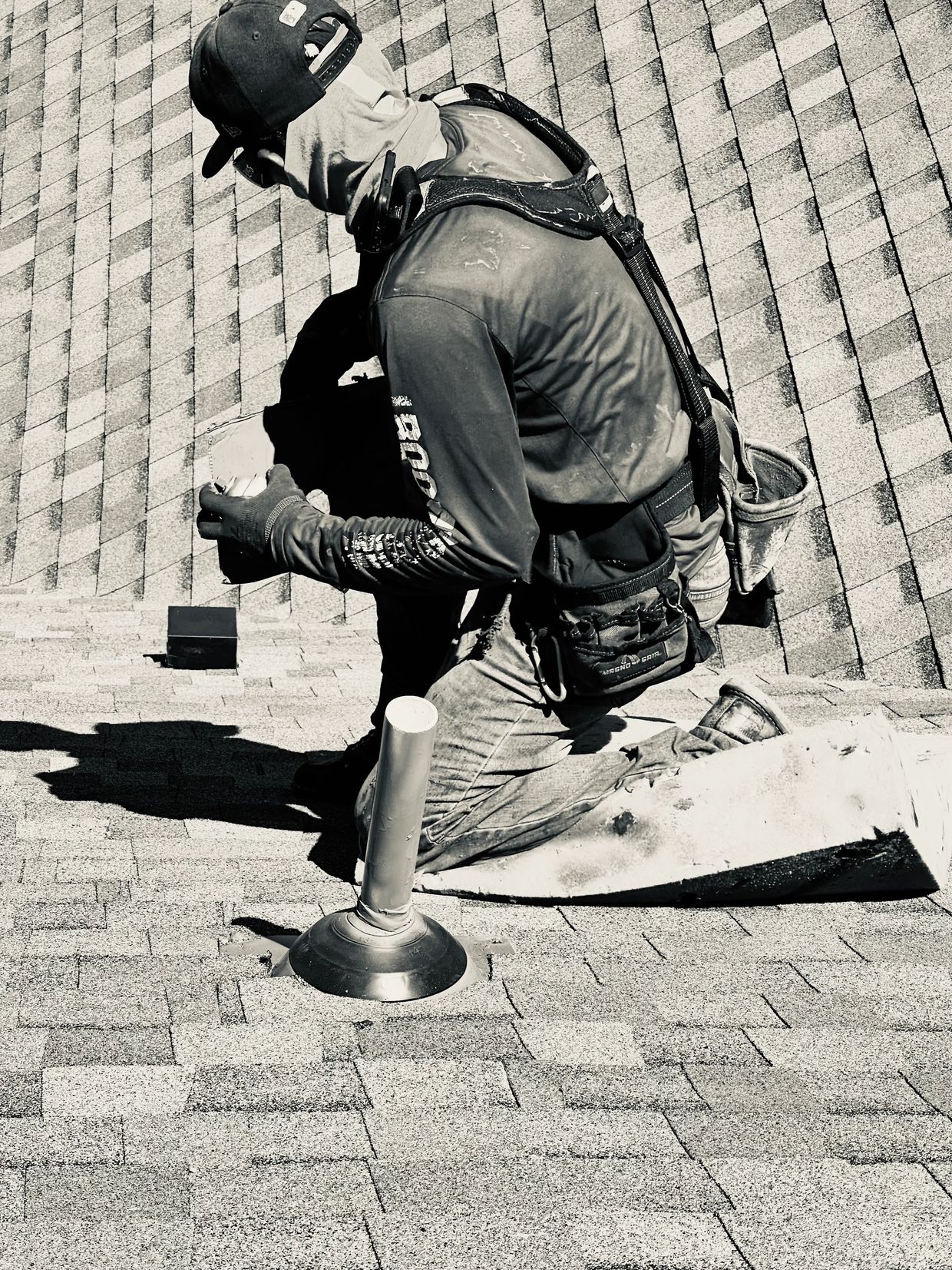 Worker kneeling on roof installing or repairing shingles with safety harness.