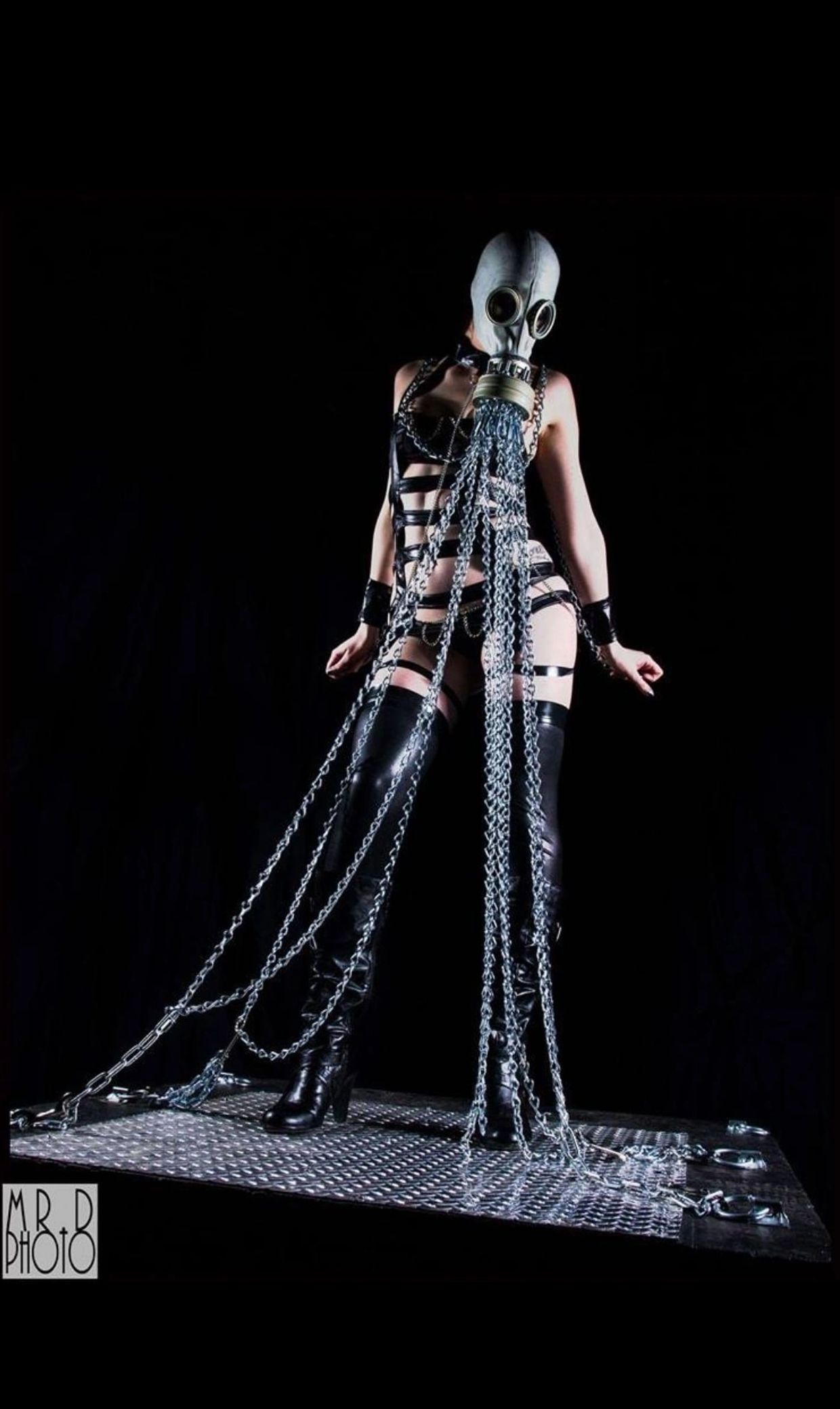 Person in a gas mask and black leather outfit with chains on a metal platform.