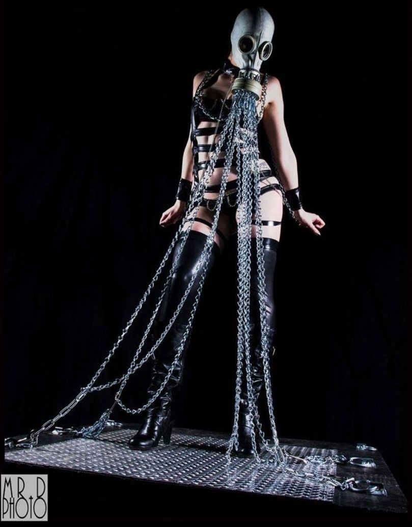 Industrial fetish photography featuring a model in strapping, boots, and a gas mask attached to cascading chains on a metal stage. Photo by Mr. D Photo.